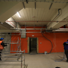 Pracownicy przenoszą rusztowania wewnątrz budynku, w tle pomarańczowa ściana