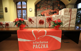 Na stole stoją zapakowane różnej wielkości paczki, z czerwonymi kokardami, przed nimi ze stołu zwisa flaga z logo Szlachetnej Paczki
