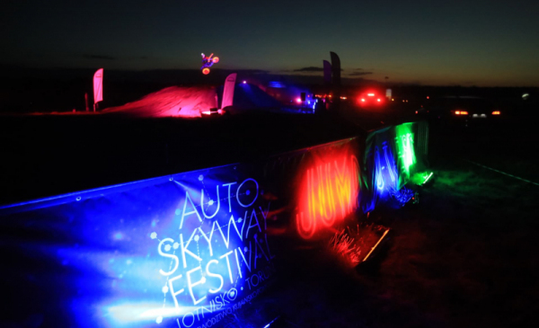 Na zdjęciu widać oświetlony fragment baneru z napisem "Auto Skyway Festival", w tle widać podświetlonego zawodnika motocrossu wykonującego ewolucję na motorze