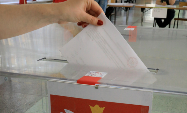 Na zdjęcli: dłoń o czerwonych paznokciach trzyma kartę do głosowania i wrzuca ją do urny wyborczej, na której jest godło kraju