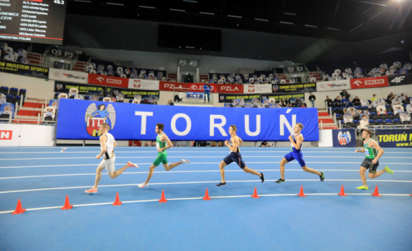 Na zdjęciu: zawodnicy biegną po bieżni w Arenie Toruń, w tle duży napis "Toruń" i herb miasta
