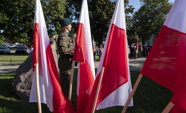 Na zdjęciu na pierwszym planie biało-czerwone flagi, pomiędzy nimi widać żołnierzy pełniących wartę przy obelisku