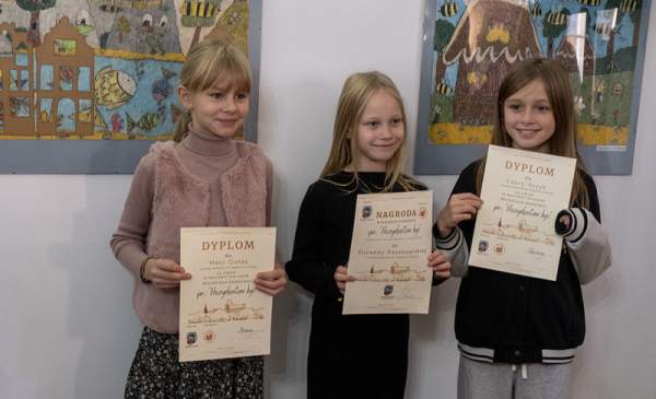 Na zdjęciu: trzy dziewczynki pokazują dyplomy, stojąc przy pracach plastycznych