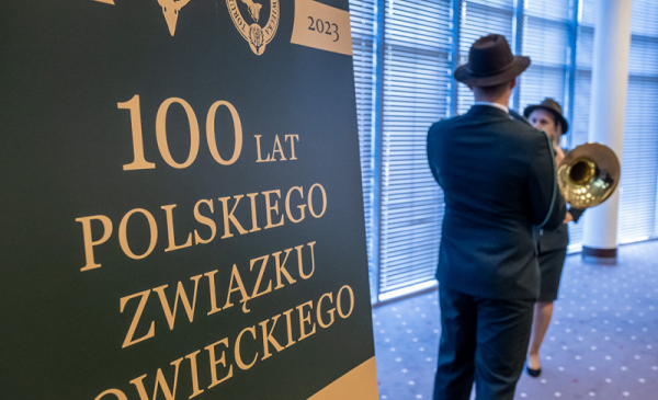 Na zdjęciu: obok  rollupu informującego o 100-leciu Polskiego Związku Łowieckiego stoi Łowczy w tradycyjnym stroju w kolorze ciemnozielonym i gra na trąbce
