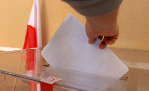 Na zdjęciu: dłoń, która trzyma kartki i wrzuca je do urny wyborczej, obok stoi flaga biało-czerwoa