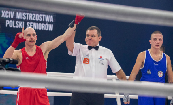 Na zdjęciu: po walce bokserskiej, sędzia podnosi rękę zawodnika w czerwonym stroju w geście zwycięstwa, stoją na ringu