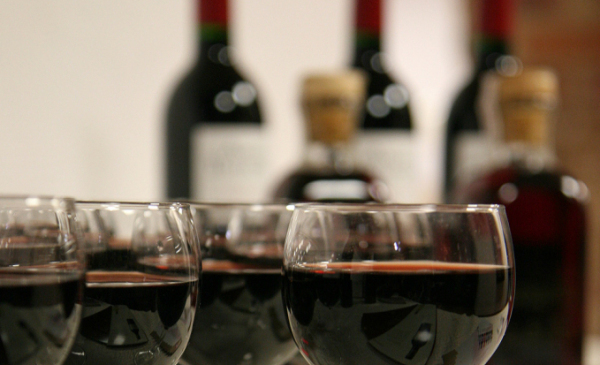 Na zdjęciu widać kieliszki napełnione czerwonym winem, w tle trzy butelki wina