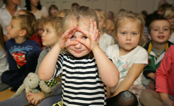 ziewczynka w przedszkolu patrząca przez dłonie zwinięte w okulary na tle grupy przedszkolaków