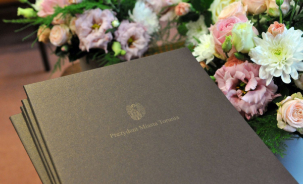 Na zdjęciu widać ciemnobrązową teczkę ze złotym tłoczeniem napisu "Prezydent Miasta Torunia", wokół leżą kwiaty