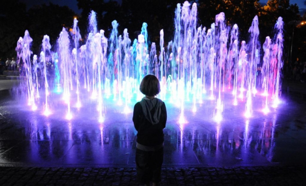 Na zdjęciu dziecko od tyłu wpatruje się w podświetloną na niebiesko i fioletowo fontannę 