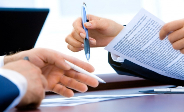 Na zdjęciu osoba trzyma długopis i kartkę papieru, druga wskazuje na dokument, który leży na biurku