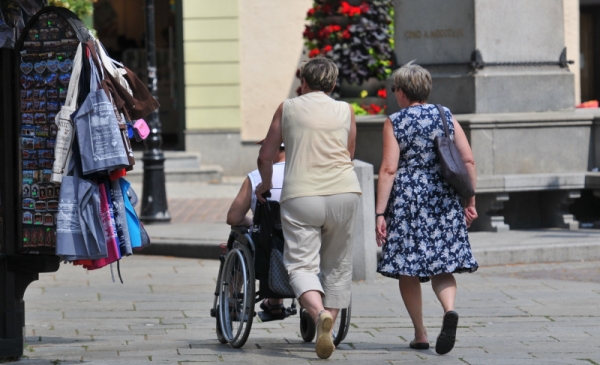 Dwie kobiety idące z wózkiem inwalidzkim pod pomnikiem Kopernika w Toruniu