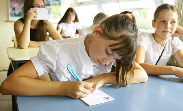 Uczennica w białej bluzce siedzi w ławce szkolnej i zapisuje coś na kartce