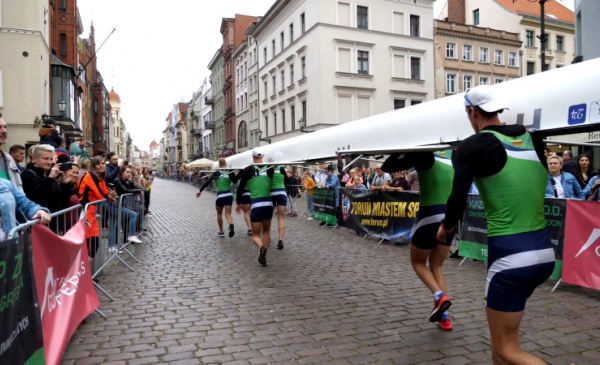 Na zdjęciu zawodnicy biegną z łodzią ulicą Szeroką, obok dopingują ich kibice