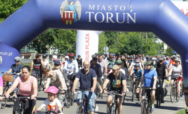 Na zdjęciu: ludzie jadą na rowerach, nad nimi duży napis Miasto Toruń i herb Torunia