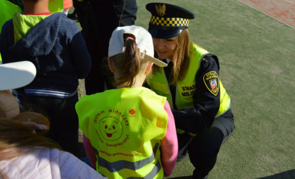 Na zdjęciu: strażniczka miejska kuca przed małą dziewczynką ubraną w kamizelkę odblaskową