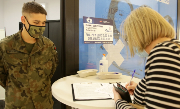 Na zdjęciu kobieta wypełnia formularz szczepienny, obok stoi żołnierz WOT