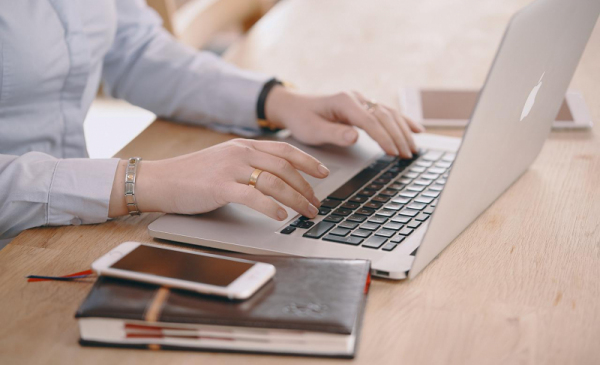 Na zdjęciu widać dłonie kobiety, pracującej na laptopie, obok leżą notes i telefon