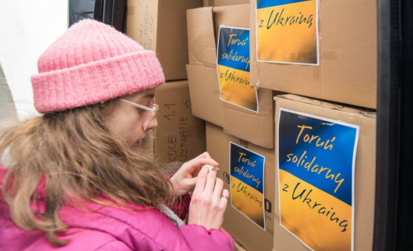 Na zdj: dziewczyna w różowej kurtce i czapce przykleja plakat z flagą Ukrainy i napisem Solidarni z Ukrainą na kartony z artykułami pierwszej potrzeby