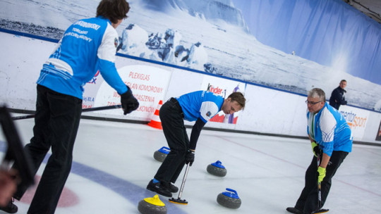 Trzech curlerów podczas meczu na torze lodowym. Niebiesko białe koszulki i czarne spodnie. Niebieskie i żółte kamienie curlingowe.