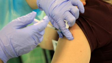 Na zdjęciu widać dłonie pielęgniarki w fioletowych rękawiczkach podczas wykonywaniu zabiegu szczepienia - igła wkłuwana jest w ramię pacjenta