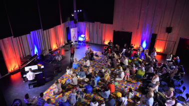 dzieci i rodzice siedzące na kolorowym dywanie w sali prób orkiestry, z przodu grają muzycy na instrumentach