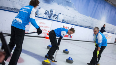 Trzech curlerów podczas meczu na torze lodowym. Niebiesko białe koszulki i czarne spodnie. Niebieskie i żółte kamienie curlingowe.