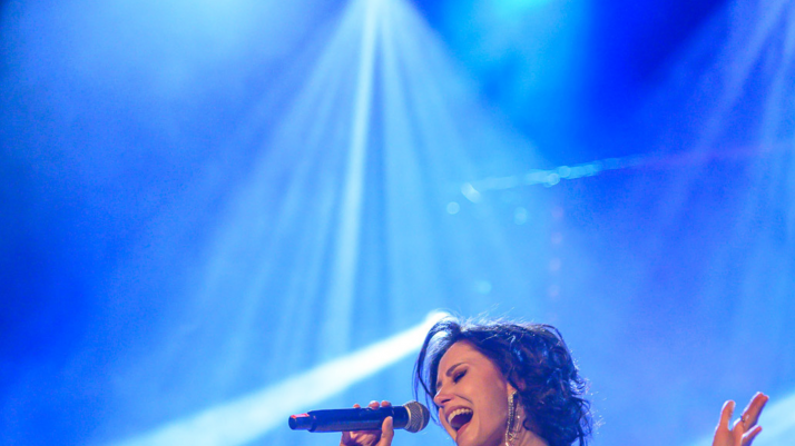 młoda kobieta śpiewająca do mikrofonu, ma szeroko otwarte usta, jasną sukienkę i ciemne włosy, w tle są światła