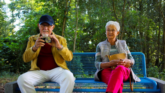 Na zdjęciu: para starszych osób, mężczyzna i kobieta, siedzą na ławce, w tle liście drzew
