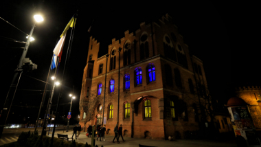 Na zdjęciu budynek urzędu miasta i okna podświetlone na niebiesko-żółto