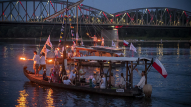 Na zdjęciu osoby trzymają pochodnie na płynącym statku, w tle most 