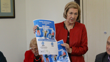 Na zdjęciu: starsza pani w czerwonym żakiecie pokazuje plakaty informujące o Radzie Seniorów
