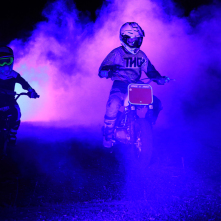 Dwóch zawodników motocrossu, oświetlonych na fioletowo, wjeżdża na teren festiwalu Auto Skyway, a wokół nich unosi się dym