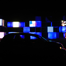 Na zdjęciu widać instalację złożoną z oświetlonych w różne wzory sześcianów, na ich tle stoi samochód osobowy