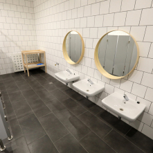 Łazienka w żłobku, szra podłoga, białe ściany z niskimi umywalkami i okrągłymi lustramiw drewnianej ramie