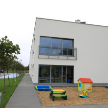 Budynek żłobka, na pierwszym planie fragment placu zabaw z plastikowym zielonym domkiem
