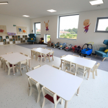 Sala w żłobku, na pierwszym planie niskie białe stoliki i krzesełka, przy ścianie stoją regały na zabawki, pod dużymi oknami leżaczki dla niemowląt, wózeczki dla lalek oraz jeździki