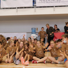 Na zdjęciu: w sali sportowej siedzą na podłodze dzieci w brązowych koszulkach