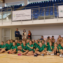 Na zdjęciu: w sali sportowej siedzą na podłodze dzieci w zielonych koszulkach