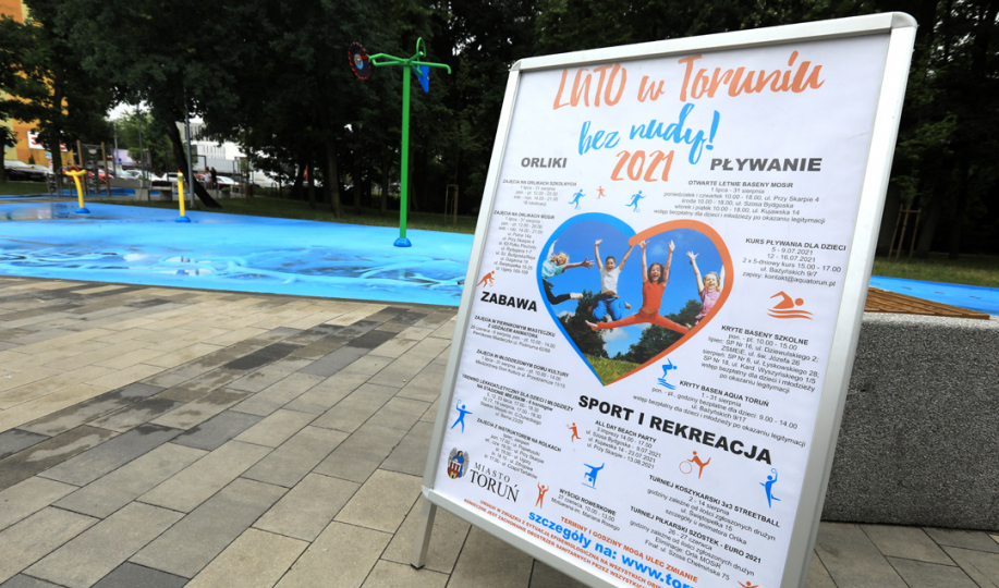 Plakat z informacjami na temat akcji Lato bez Nudy ustawiony przy wodnym placu zabaw przy Aqua Toruń