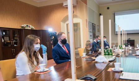 Pięcioro absolwentów siedzi przy stole, od pozostałych uczestnikow spotkania oddziela ich plastikowa ścianka