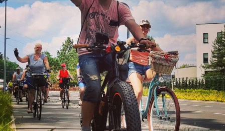 Mężczyzna z brodą w różowej koszulce i czapczce pozdrawia fotografa z roweru
