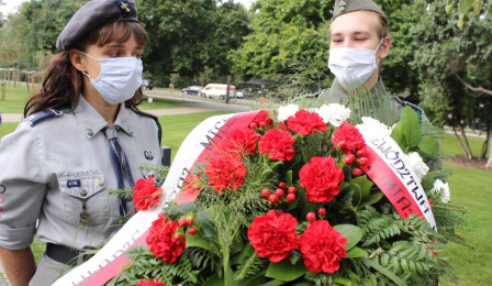 Na zdjęciu widać wiązankę kwiatów z biało-czerwoną szarfą, w tle stoi dwoje harcerzy